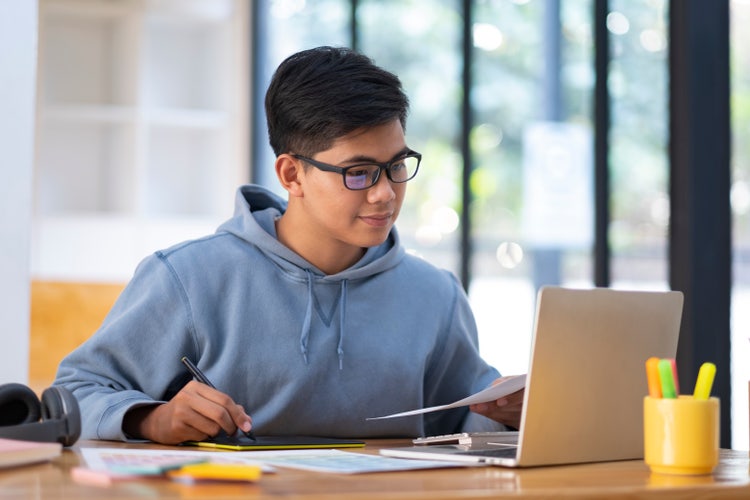 A man in a blue hoodie sits at a desk with a piece of paper in his left hand while looking at his laptop and writing a synthesis essay.