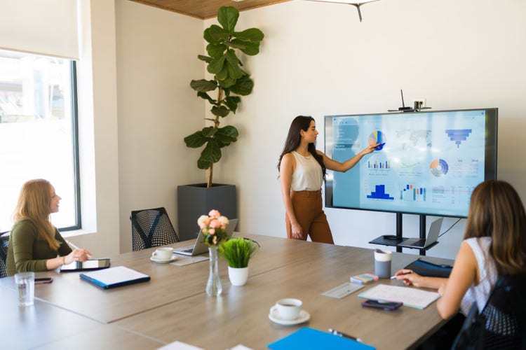 Two women sitting at a large desk watch another woman give a presentation using an ODP file on a large monitor.