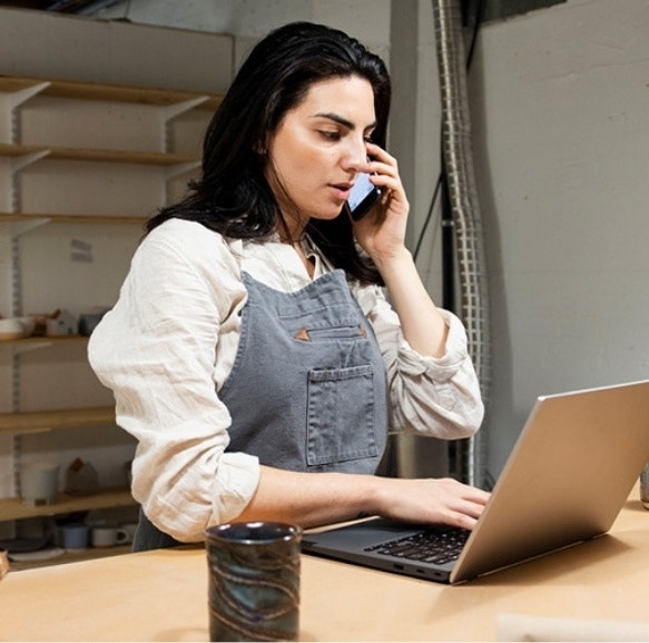 A person in overalls talking on the phone while on a laptop discussing details of their pottery business.