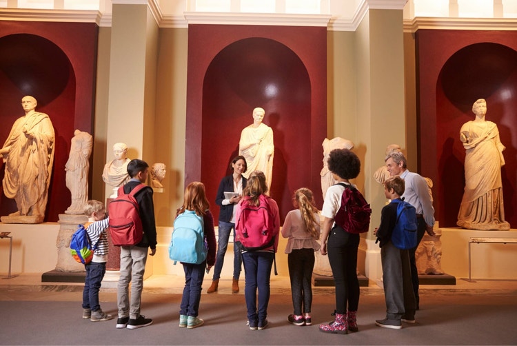 A museum guide giving a tour to a group of students and their teacher in front of marble statues