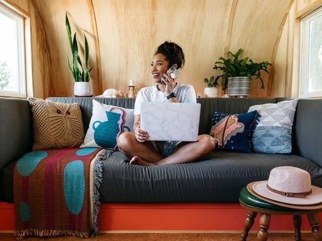 A person sitting on the couch of a converted and decorated airstream trailer chatting on the phone while multitasking on their computer