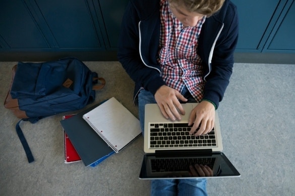 A photo of a student sitting against lockers on the floor of a hallway and working on a laptop.