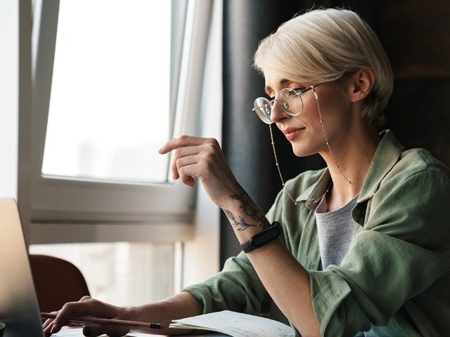 A fashionable person with wireframe glasses working on a notebook with pencil in hand while also typing on a laptop near a window