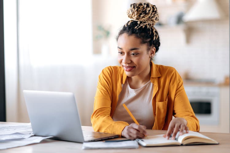 A photo of a person sitting at a table in a home, looking at a laptop, and taking notes with a pen on notepad.