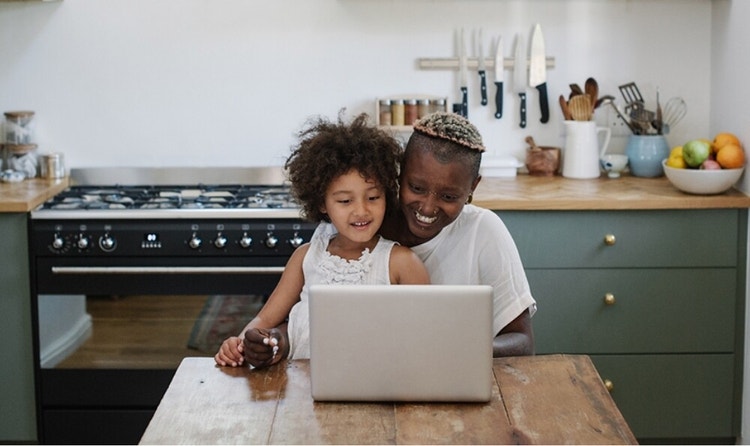 A young girl sitting on her mother's lap at a reclaimed wood table as they both smile and look over something on a laptop
