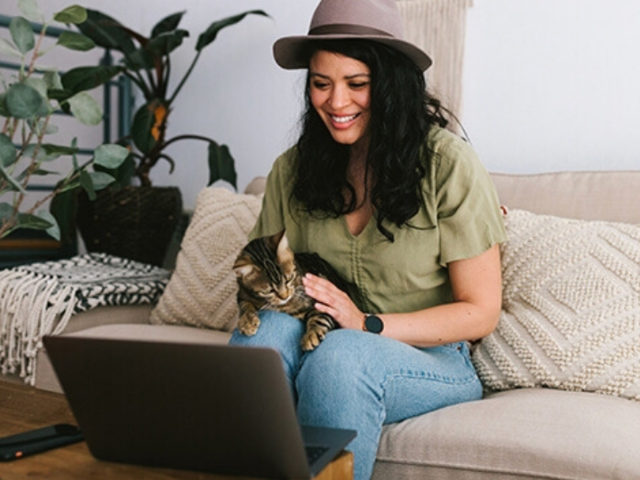 A person working remotely on their laptop while sitting on their couch with a cat in their lap