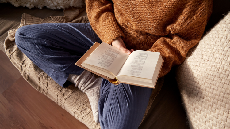 A person sits cross-legged on a sofa with an open book in their lap