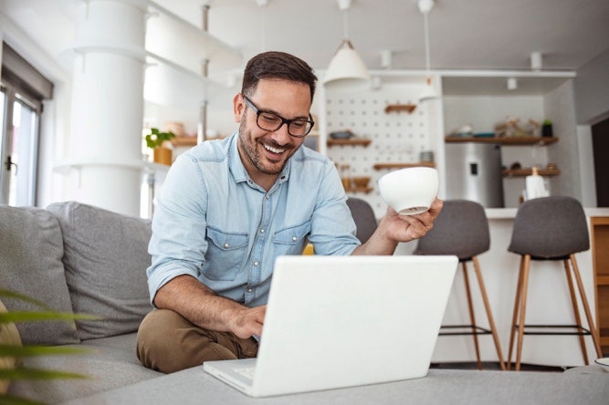 A person using their laptop while enjoying a cup of coffee and sitting on a sofa.