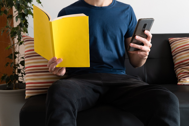Male sitting on a sofa holding both a paper notebook and a mobile phone.