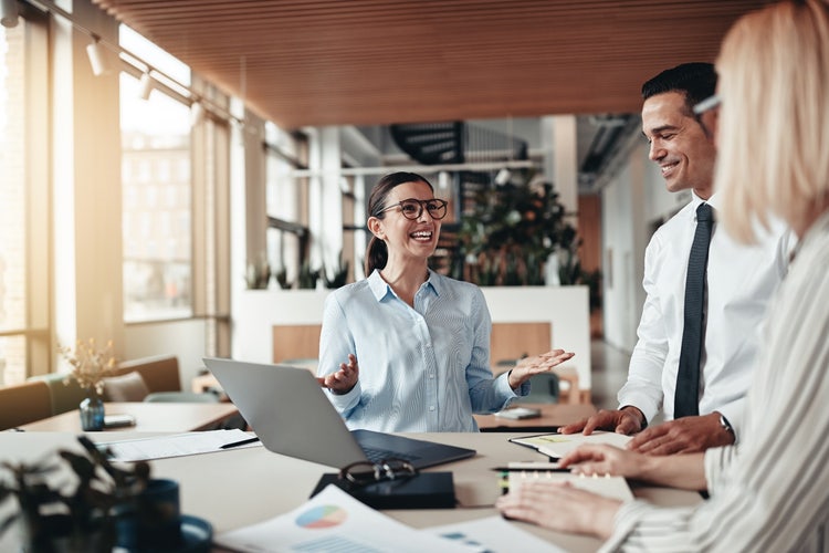 Two women and a man are chatting at a desk with a laptop and various office stationery surrounding them