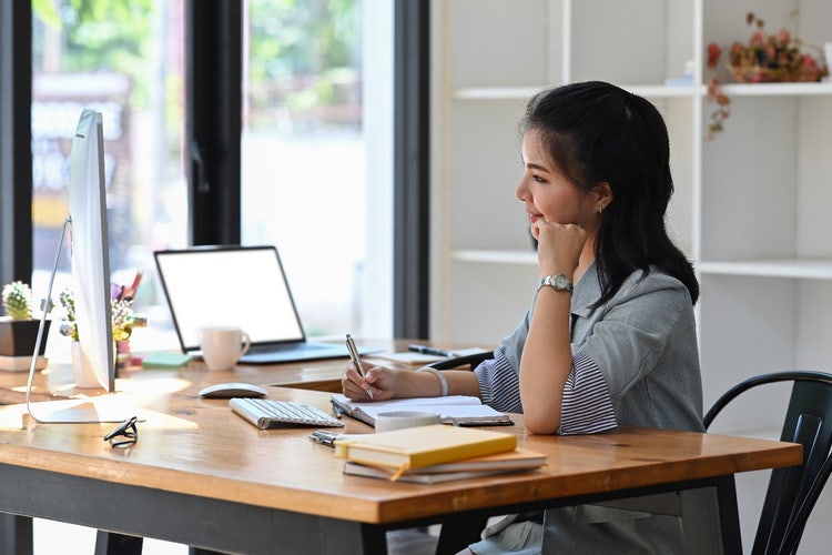 Woman sitting at a desk reading a computer screen with a notepad and pen in hand.