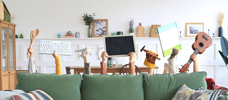 Photo of children's arms raised behind a couch and in front of a dining table. They children are holding up items such as a keyboard, chalkboard with Adobe written on it, wooden spoons, hammer, book, and musical instruments.