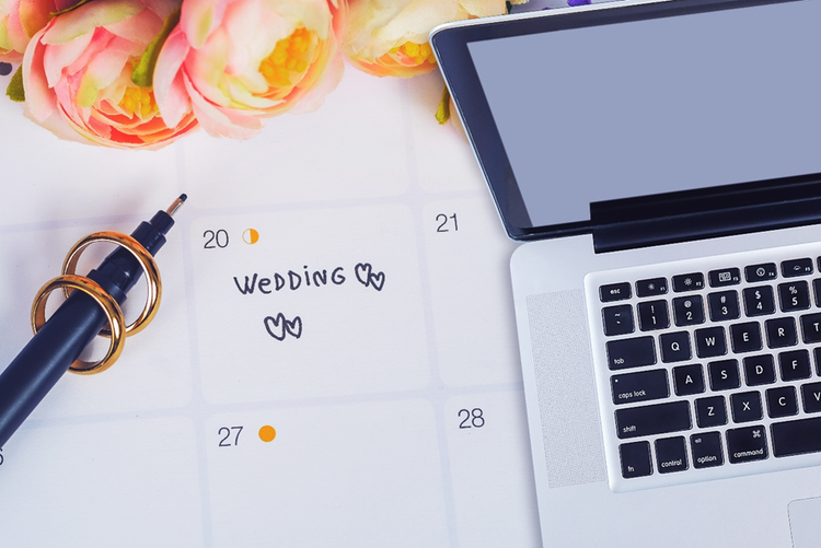 Photo of a laptop on top of a calendar the word "wedding" and hearts drawn on the date. Two rings have been slide over the pen and fresh flowers are at the top of the calendar.