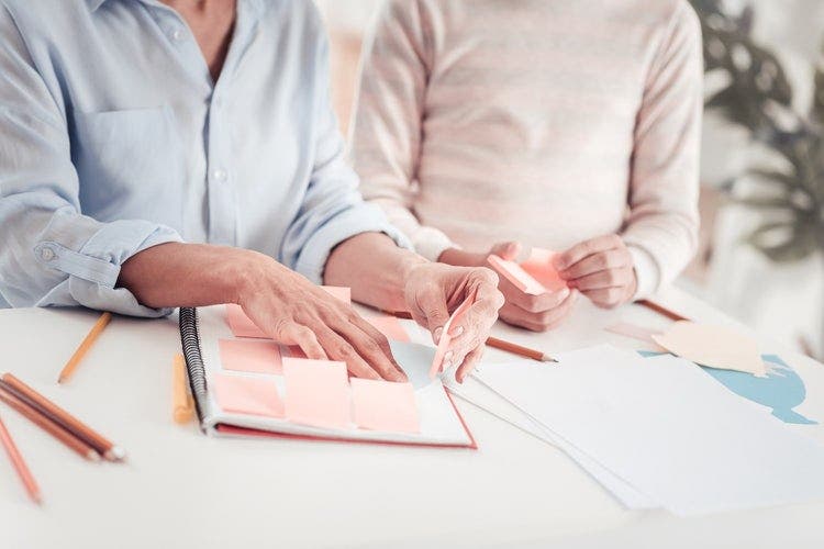 Photo of a woman and child sitting at a table with a notebook and pencils, and placing sticky post-it notes on the paper pages of the notebook.