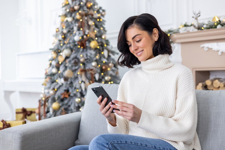 Photo of a woman sitting on a sofa near a decorated Christmas tree, smiling as she edits photos on her mobile phone.