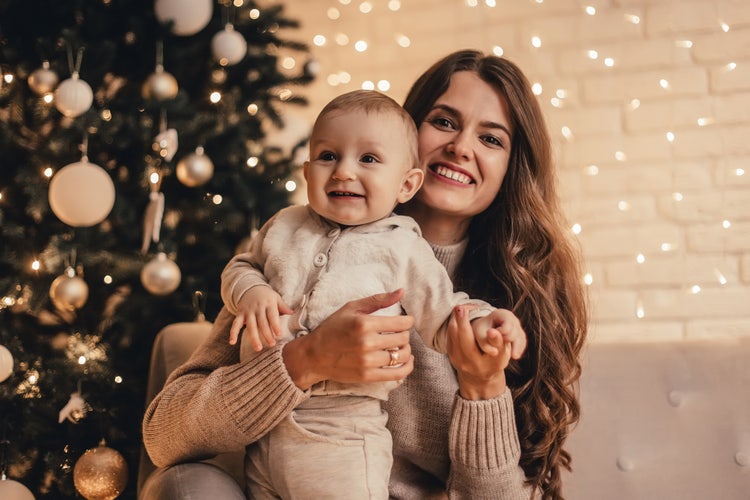 Happy mother and her young child hugging and celebrating the holidays in front of a decorated Christmas tree.