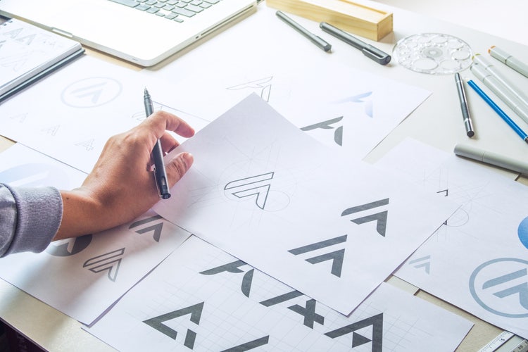 Designer sketching brand logo concepts on paper, with tools and a laptop on the desk.