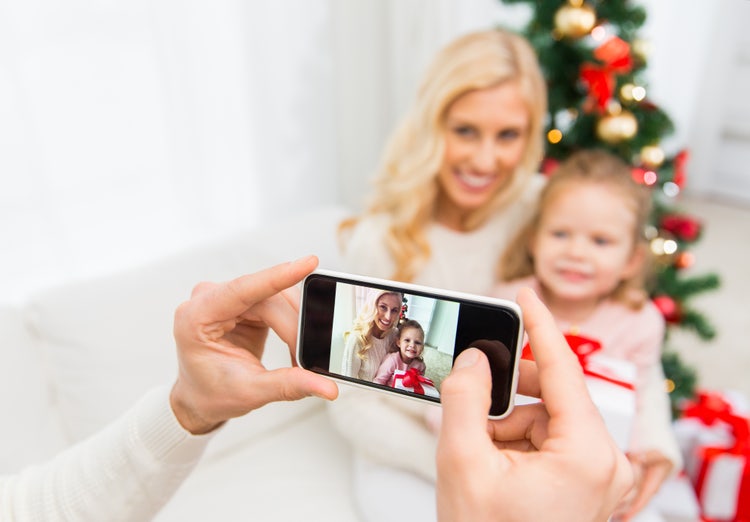 A person is taking a photo of a mother and child smiling together in front of a Christmas tree, with gifts and decorations in the background.