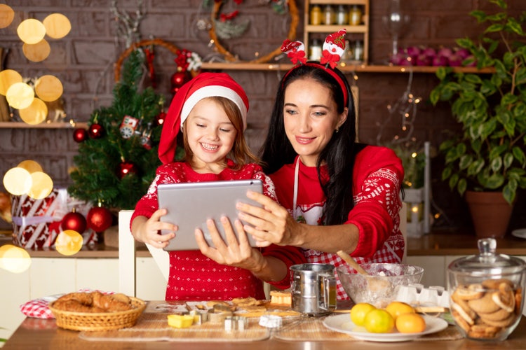 Mother and daughter in festive outfits, smiling and looking at a tablet together while baking Christmas cookies in a decorated kitchen with a Christmas tree in the background.