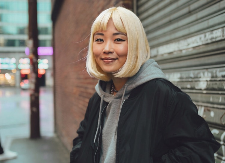 Image générée par l’IA d’une jeune femme au look décontracté et aux cheveux blond platine, dans une rue en ville devant un mur de briques et un rideau métallique