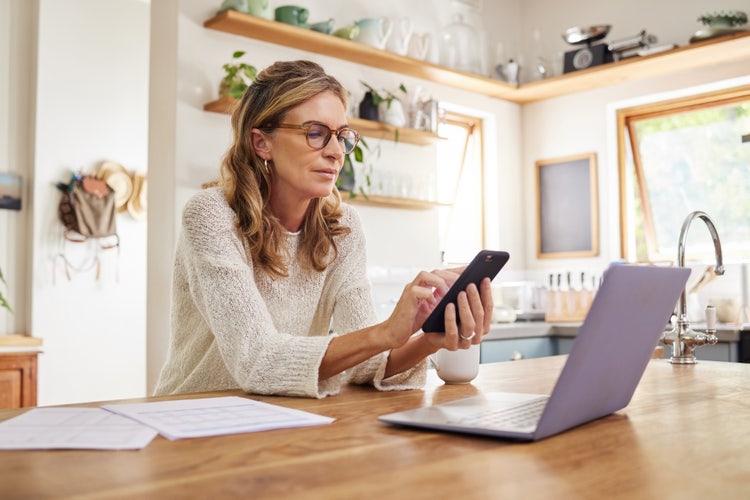 Uma mulher sentada à mesa da cozinha usando o telefone e o notebook para digitalizar documentos.