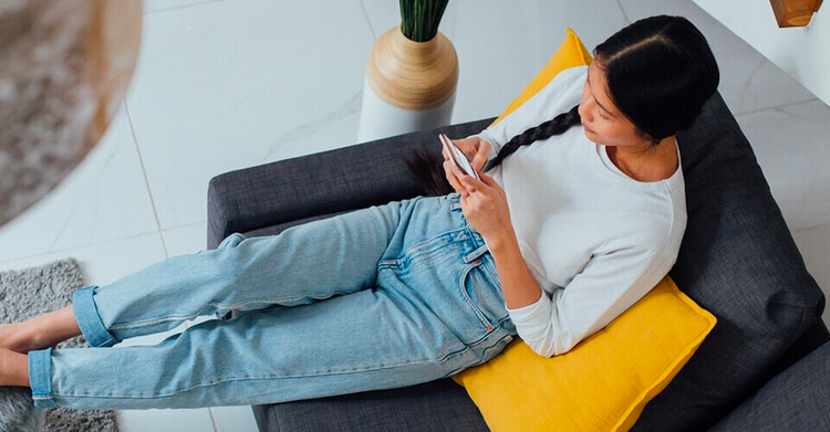 Mulher, sentada em um sofá com as pernas esticada, digita em um smartphone