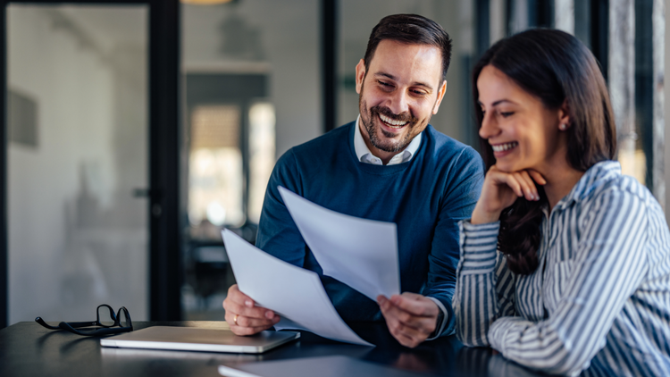 A man and a woman sit at a table and smile as they look at a few pages of paper.