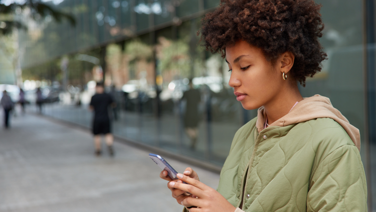 A woman stands outside looking at her phone. She is wearing a green jacket and gold hoop earrings.