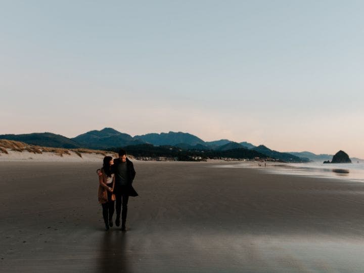 A long shot of a couple in casual attire walking along a sandy beach at dusk are captured in a long shot