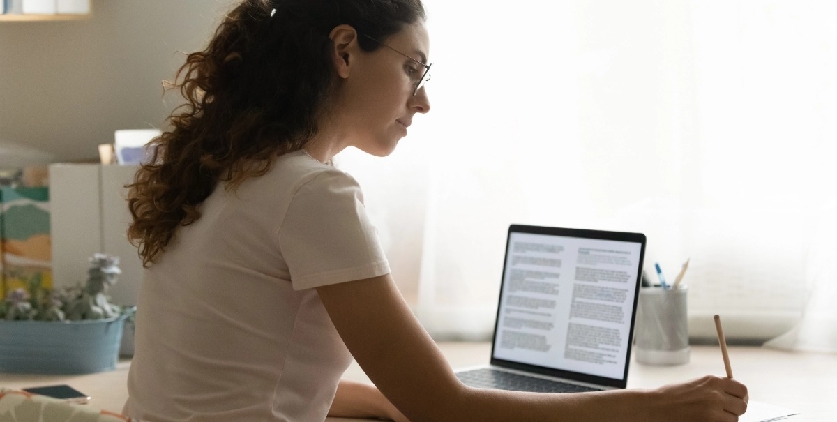 Foto: Mujer tomando notas, junto a un portátil con un documento de texto abierto.