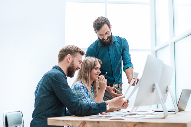 Three businesspeople compare notes happily in front of three computer monitors.