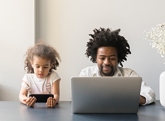 A parent using their laptop and their child looking at their own phone while sitting at a table together