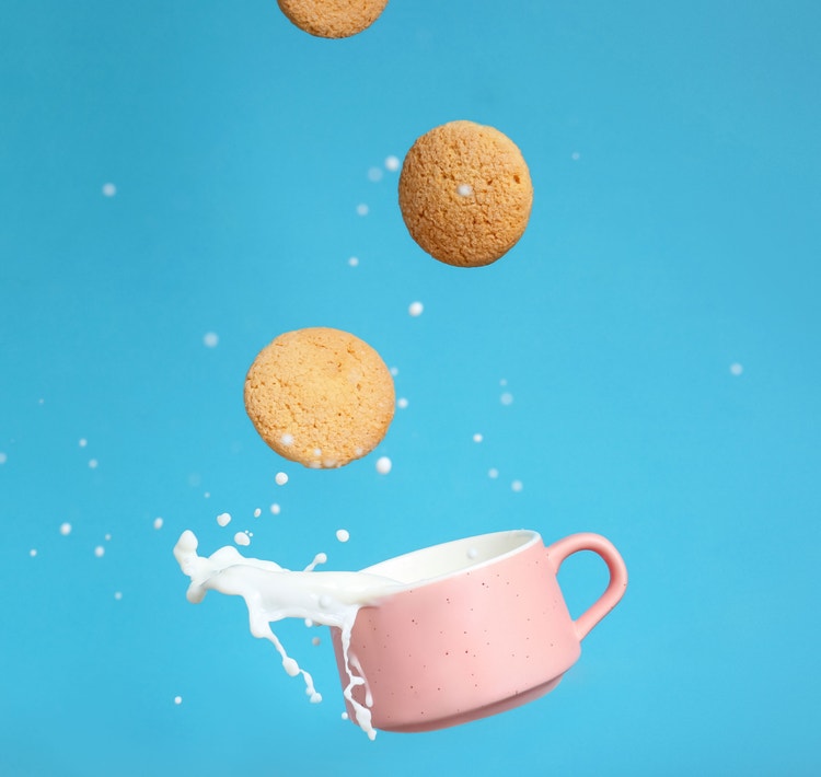 Cookies falling into a pink mug and splashing milk on a blue background.