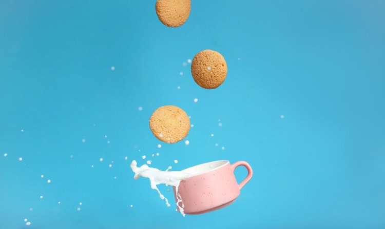 Cookies falling into a pink mug and splashing milk on a blue background.