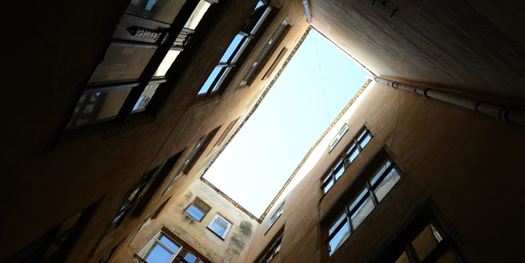 A low-angle shot depicts looking up into a rectangular blue sky between four dark brown building walls with windows.