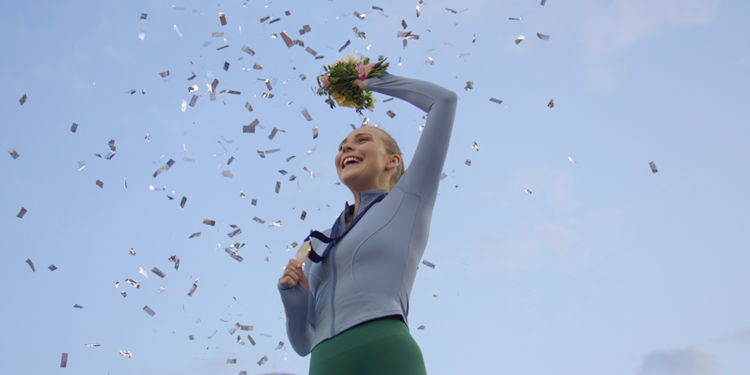 A low-angle shot of a woman with a gray long-sleeved running shirt throwing confetti against a clear blue sky.