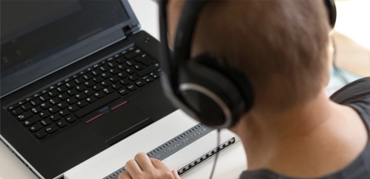 Foto einer Person mit Kopfhörern vor einem Laptop mit Blindenschrift.