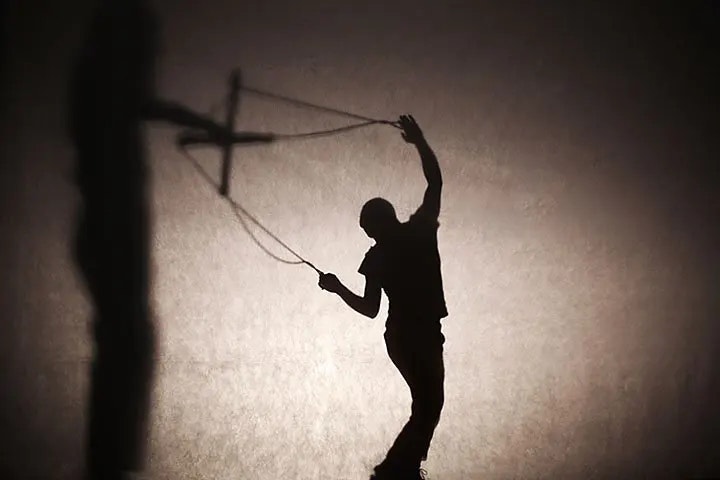 Schattenumriss eines Puppenspielers, der eine Marionette an Fäden steuert. Die Szene ist in Schwarz-weiß gehalten. Die Schatten des Puppenspielers und der Marionette sind vor einer hellen Leinwand projiziert.