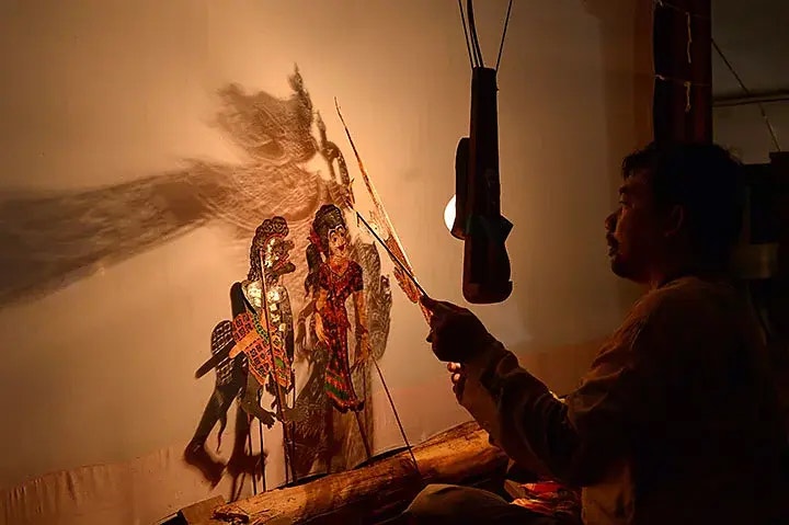 Ein Puppenspieler führt ein traditionelles Schattenspiel durch, bei dem kunstvoll gestaltete Puppen vor einer beleuchteten Leinwand bewegt werden. Der Puppenspieler ist seitlich zu sehen, während die Schatten der Figuren auf die Leinwand projiziert werden.