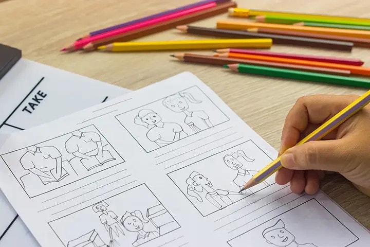 Hand zeichnet einfache Comic-Panels auf einem Blatt Papier, umgeben von bunten Bleistiften auf einem Tisch. Die Skizzen zeigen verschiedene Figuren in verschiedenen Szenen.