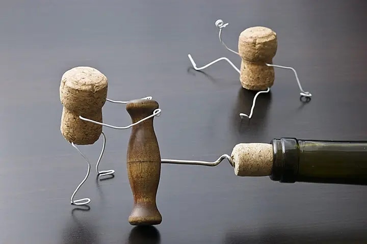 Zwei Korken mit Drahtarmen und -beinen sind wie Figuren gestaltet. Eine hält einen Korkenzieher, der in eine Weinflasche gedreht ist. Der andere sitzt im Hintergrund.