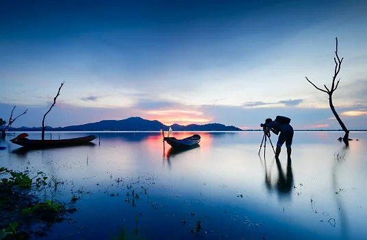 Ein Fotograf steht in seichtem Wasser und fotografiert einen Sonnenuntergang hinter einem Gebirgszug