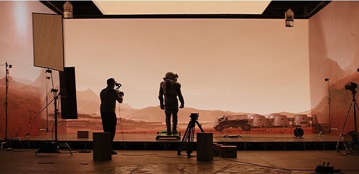 Blick auf ein Filmset im Studio. Ein Kameramann im Vordergrund filmt einen Schauspieler in einem Raumanzug vor einer futuristischen Kulisse
