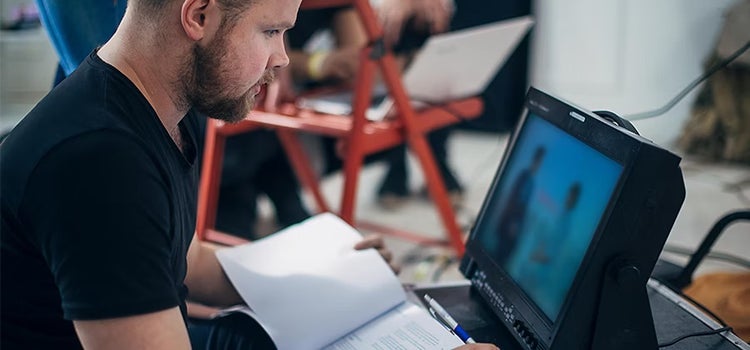 Mann links im Bild hat ein Drehbuch auf dem Schoß und beobachtet die Dreharbeiten über einen Bildschirm, der rechts im Bild ist. Im Hintergrund ist ein roter Stuhl mit einem Laptop darauf zu sehen