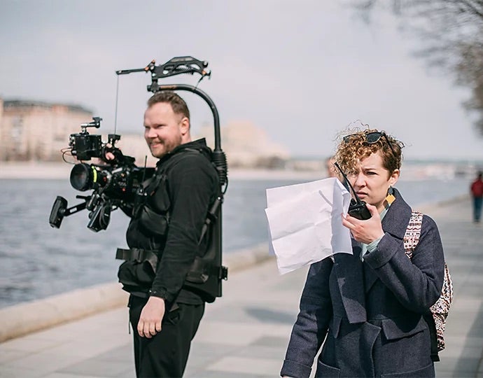 Ein Kameramann links im Bild wartet auf seinen Einsatz. Rechts neben ihm steht eine Regieassistentin mit einem Walkie-Talkie und Notizen in der Hand