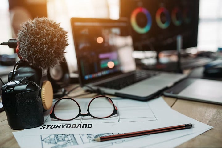 Blick auf einen Schreibtisch mit einer Kamera mit Mikrofon links, einem Laptop im Hintergrund und einer Brille und einem Bleistift, die auf Unterlagen für ein Storyboard liegen