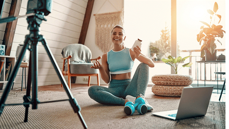 A woman sits on a carpeted floor with two objects in her hands. She smiles at a camera on a tripod and has a laptop by her feet.