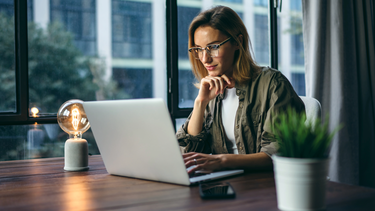 A woman sits at a table and looks at her laptop. She is wearing glasses and a green shirt.