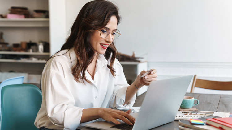 A brown haired woman looks at her open laptop while seated at a table. She is wearing glasses.