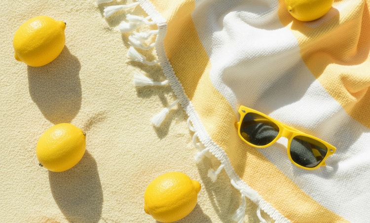Sommerliches Flat-Lay von oben mit leuchtend gelben Zitronen und passender Sonnenbrille auf einem gestreiften Strandtuch neben hellem Sand, beleuchtet von warmem Sonnenlicht.